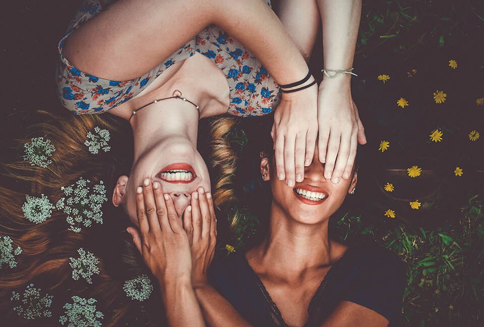 Two women laying in the grass covering each other's eyes and laughing