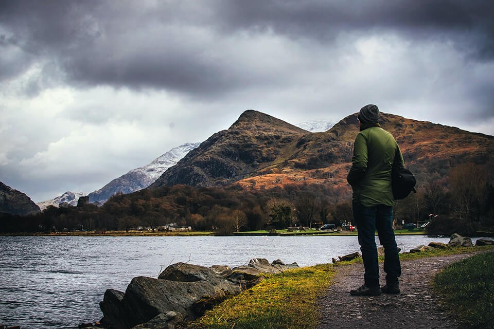 Man standing on path, looking over a cloudy sky and peaceful lake.