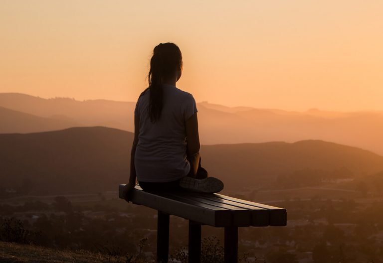 a woman sitting on a bench looking at the sunset.