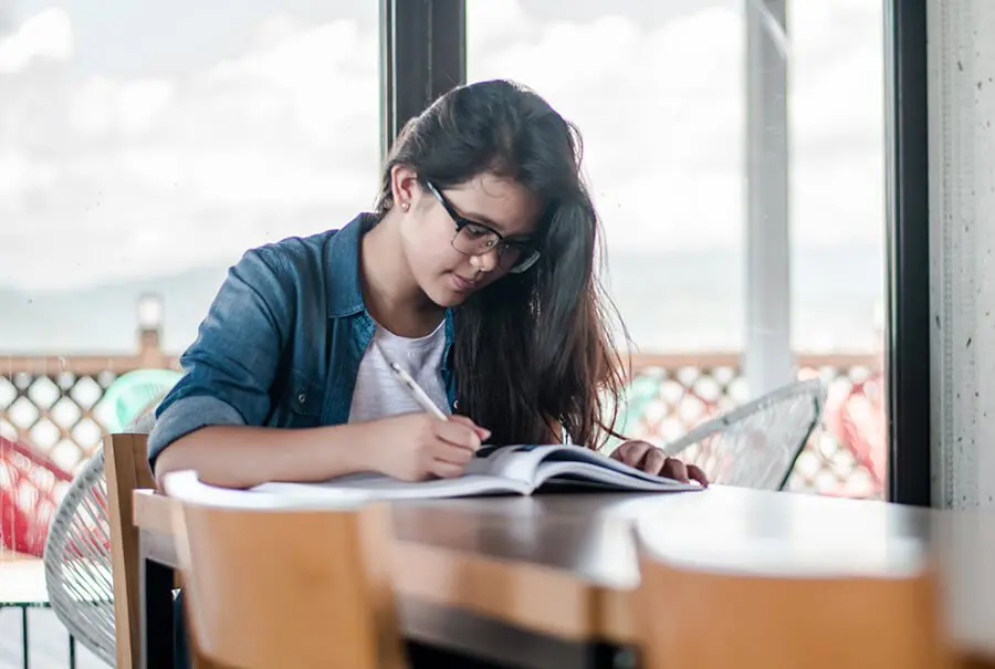 a girl is sitting at a table writing on a book.