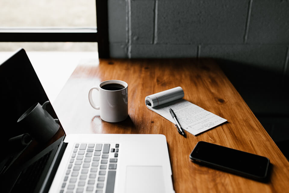 Laptop opened on desk with coffee and pen and pad of paper